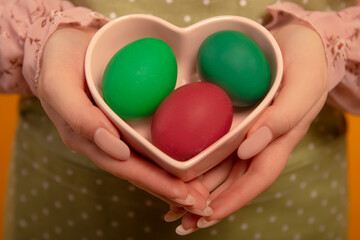 Easter day concept. Close-up view of female hands holding heart shaped plate saucer with two green dyed easter eggs and one red painted easter egg isolated on green polka dot kitchen apron.