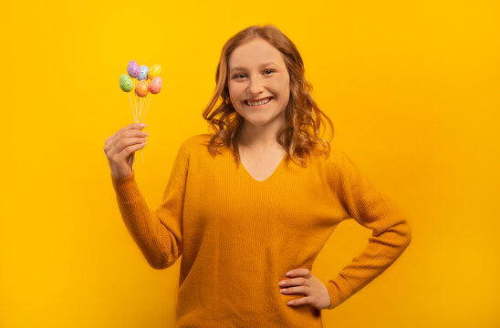 Happy smiling laughing young woman in yellow sweater holding small painted dyed easter eggs on sticks, keeping hand on waist hip isolated on yellow background.

Easter Day celebration concept.