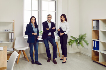 Successful business team standing together looking at camera and smiling. Portrait of smiling business people in formal wear standing near window in modern office. Collaboration, teamwork, partnership