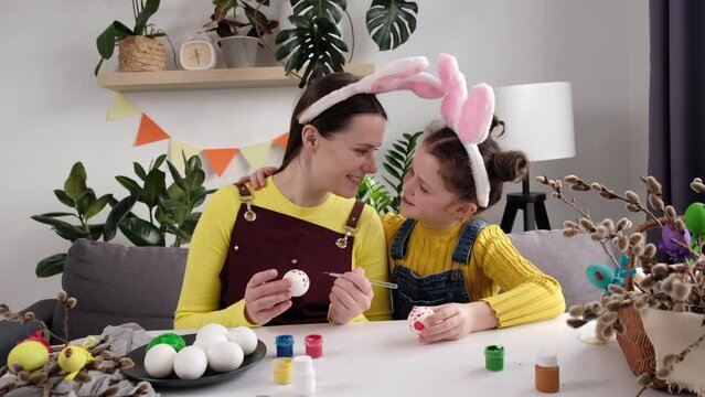 Loving sweet little daughter gently cuddles kiss mother on cheek during painting eggs sitting together at table. Happy family preparing for Easter. Pretty little girl child and mom wearing bunny ears