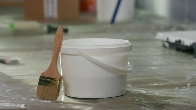 A round plastic bucket with a handle and brush stand on the floor covered with oilcloth. Close up of a white pail with paint and a clean wooden brush against a blurred room background.