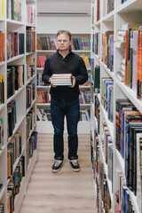 young adult man stands in library in middle of the shelves and holds stack of books in hands. education at university campus, free training in exact sciences. bookstore. successful male student 
