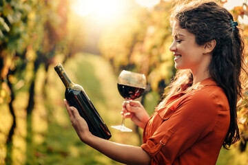 Woman holding glass and bottle of red wine in vineyard - Powered by Adobe