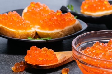 Red caviar close-ups on wooden spoon, in glass plate and on buns with butter and parsley on black plates on gray background.