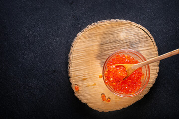 Red caviar in wooden spoon on glass bowl that stands on log from tree on black background. Wooden spoon with glass cup filled with caviar stand on log on black background.