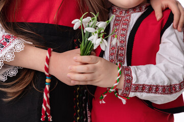 Fototapeta na wymiar Bulgarian kids boy and girl in traditional folklore costumes with snowdrop flowers and handcraft wool bracelet martenitsa symbol of 1 March Baba Marta, spring and Easter, martisor