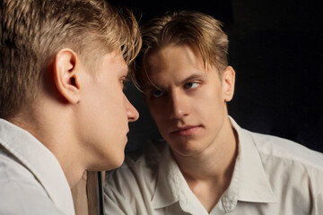 a serious man in a white shirt looks at himself in the mirror standing on a black background