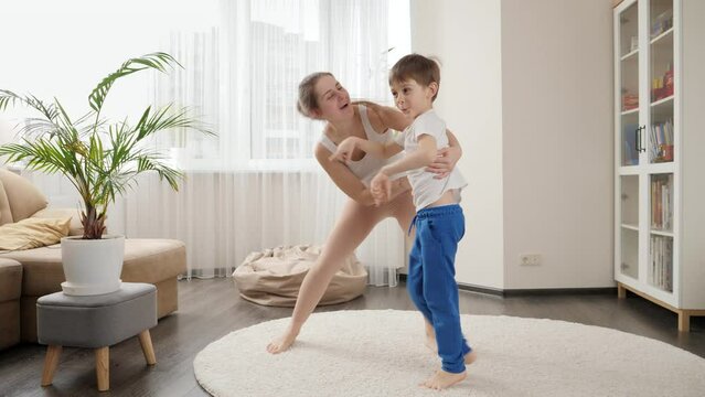 Funny shot of happy smiling mother playing and dancing with her little son in living room at home. Family having fun together, listening music, active lifestyle, parenting and child development.