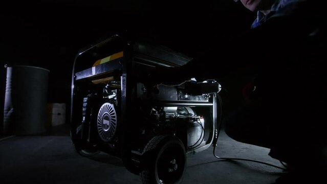 A man in blue overalls and a yellow helmet in the dark turns on a gasoline generator and lights up. The worker puts on goggles and cuts metal with a grinder or a circular knife, sparks fly.