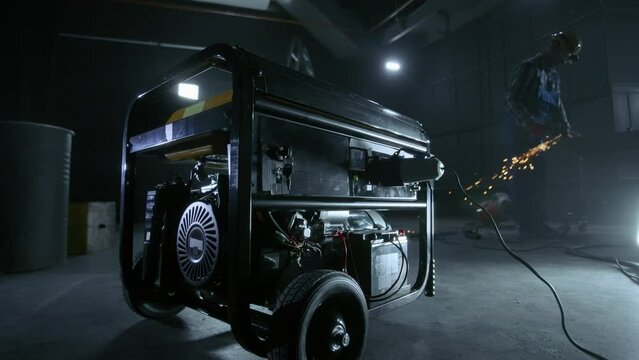 Black mobile gasoline power generator in a dark workshop. In the background, a man in blue overalls, a hard hat and goggles cuts metal with a grinder or a circular knife. In the process, sparks fly.