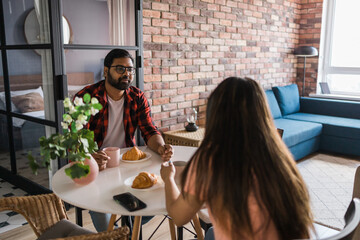 Young diverse loving couple eating croissant and talks together at home in breakfast time. Communication and relationship concept