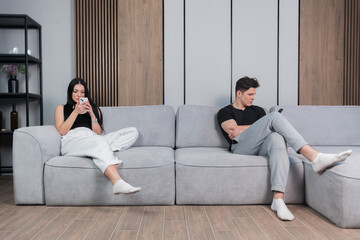 A man and a woman sitting on a couch ignore each other and use their cellphones as they are both uninterested in their relaationships. family problems. Couples with trust issues.