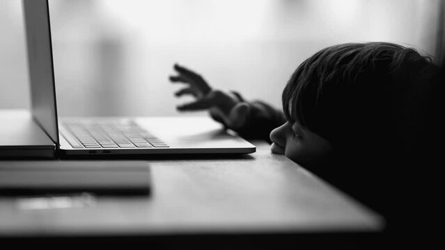 One bored little boy leaning on table and using computer laptop feeling boredom in monochromatic black and white