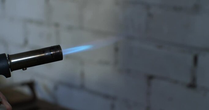 A man's hand holds and turns on a blowtorch. The blowtorch flashes brightly after being turned on and burns with a steady blue flame.
