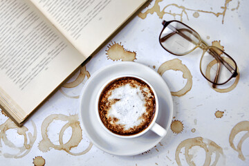 A cup of coffee beverage, reading glasses and a paper old book on a white table with coffee stains