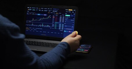 A trader spins bitcoin in his hand while looking at the chart of cryptocurrency growth on the spot. A trader counts dollars in front of a laptop screen with a cryptocurrency candlestick chart.