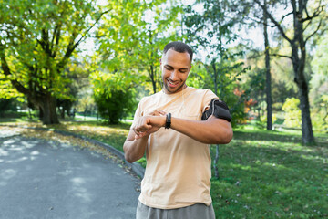 Fototapeta na wymiar A young male African American athlete is standing in the park and going for a run. Adjusts a smart watch, fitness bracelet on the hand.