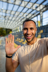 A young African American male athlete is standing in a stadium, holding a phone and talking on a video call through headphones. He looks at the camera, smiles, says hello. Vertical, close up photo.