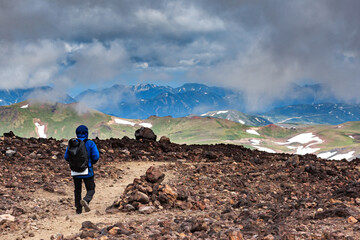 Fototapeta premium A tourist walks on volcanic sand, against the backdrop of a volcano, trekking, Kamchatka, tourist, mountains, clouds.