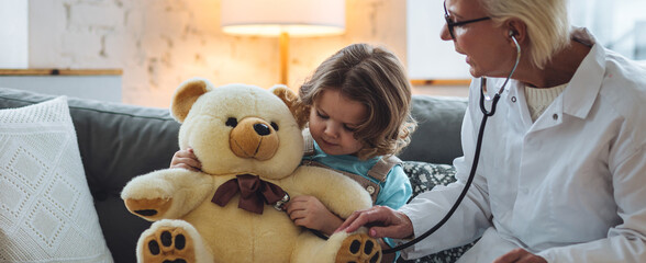 Kind female senior paediatrician doctor visiting his patient at home, examining little girl with teddy bear toy. Concept of kid's health check. Measure temperature, heart beat. Banner