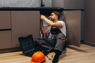 A young man fixes a pipe in the kitchen under the sink.
