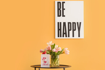 Vase with tulips and greeting card for Women's Day on table near yellow wall in room