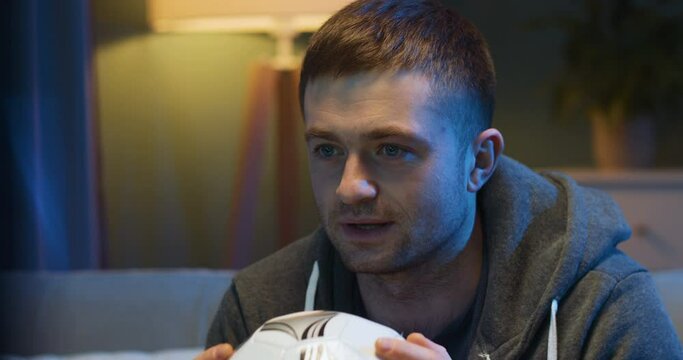 Close up of worried handsome Caucasian guy sport fan watching match on TV late in evening at home. Smiled man worrying and cheering for favorite football team during championships.