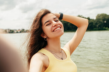 Naklejka premium Woman standing smiling near river sea lake, take picture, make selfie hold camera. Female on beach island. Girl tourist on vacation, weekend relaxing resting resort. Lady go on shore, sunbathing 