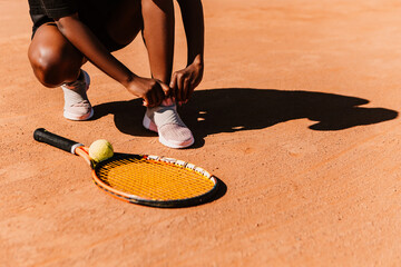 African american woman with racket and yellow green ball, preparing shoes for playing tennis match on clay court surface on sunny day. Female player ready to serve. Professional sport concept
 - obrazy, fototapety, plakaty