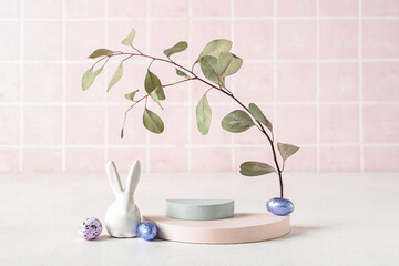 Podiums with Easter eggs, rabbit and tree branch on table near pink tile wall