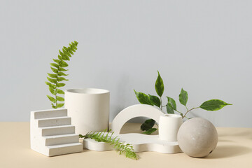 Plaster podiums with fern and ficus branches on beige table against grey background