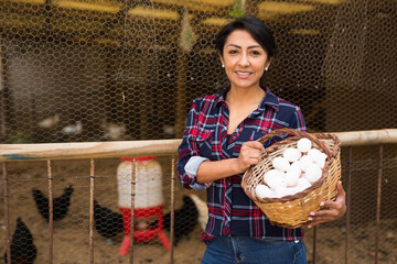 Smiling latin american woman worker holding basket of fresh organic eggs at poultry farm