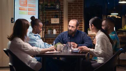 Diverse coworkers brainstorming, team leader explaining strategy plan in business meeting, discussing marketing presentation. Employees analyzing data at boardroom table, talking in coworking space