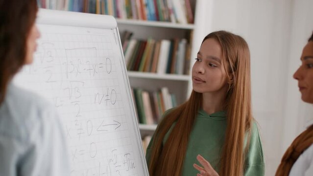 Extra classes. Group of young ladies students listening to tutor explanation, standing at whiteboard with math formulas