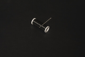 A lone spool of shiny black thread with a pricked sewing needle lying on a black surface