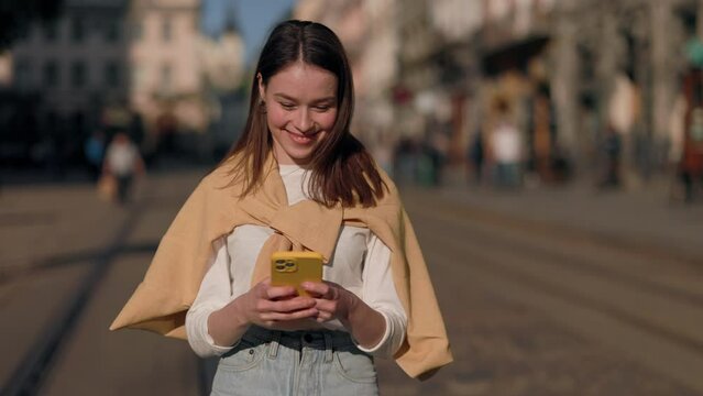 Charming caucasian brunette woman using smartphone at city walk sunset look smile portrait close up slow motion. Girl texting on gadget outside. Blur background of city street.
