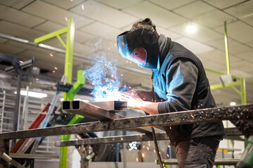 A man working as a welder in a protective mask and work clothes performs work with a MIG-MAG...