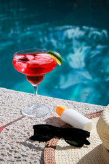 refreshing red Cocktail at the pool with sunscreen, sunglasses and straw hat
