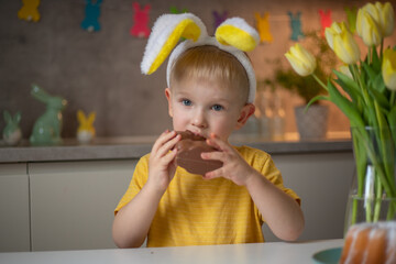 A cute little boy wearing bunny ears on Easter day is eating a chocolate Easter bunny. A child...