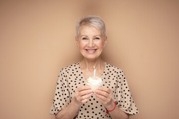 elderly woman with short haircut holds a cupcake with a candle in her hands, celebrates a birthday...