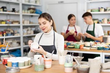 Fototapeta premium Young woman in apron paints ceramic cup in workshop