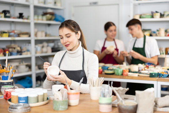 Young woman in apron paints ceramic cup in workshop