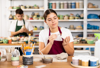 Fototapeta premium Smiling young woman in apron painting on handmade clay cup with brush in pottery workshop. Persons viewing handmade pottery products