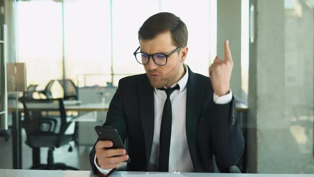 Angry boss shouts to the employee while looking at phone, dismisses the subordinate, a businessman in a business suit sits at a table in a modern office