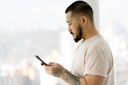 Side View Of Asian Man With Stylish Tattoos Holding Smartphone Using Mobile App Shopping Online. Bearded Hipster Guy Reading Text Message Standing Near Window At Home 