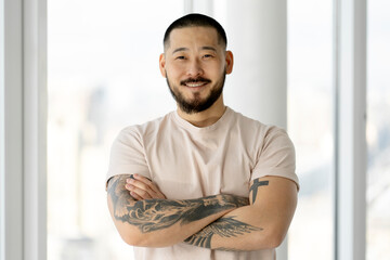 Portrait of handsome smiling Asian man with arms crossed and stylish tattoos looking at camera
