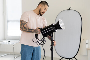 Professional Asian photographer adjusts lamp with lighting, preparing for shooting.  A young man works in a professional photo studio 