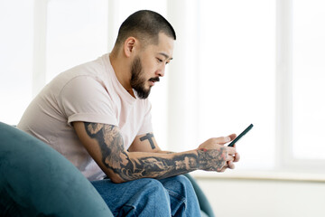 Serious handsome Asian man using mobile phone , shopping online sitting at home. Hipster guy with stylish tattoo chatting, reading text message, communication online. Technology concept