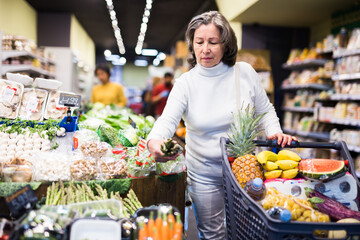 Obraz premium Focused aged woman standing with shopping trolley full of fresh produce in vegetable section of supermarket, choosing fresh green asparagus