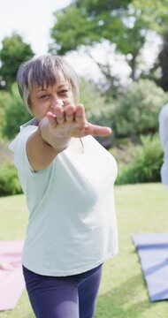 Vertical video of happy senior biracial woman doing yoga with friends in garden, in slow motion